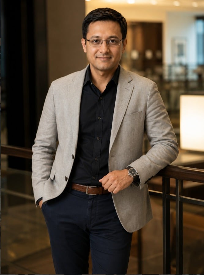 Dr. Siidarth Bhattacharya, founder of Heal & Coach, in a light grey blazer leaning on a railing in a warmly lit interior